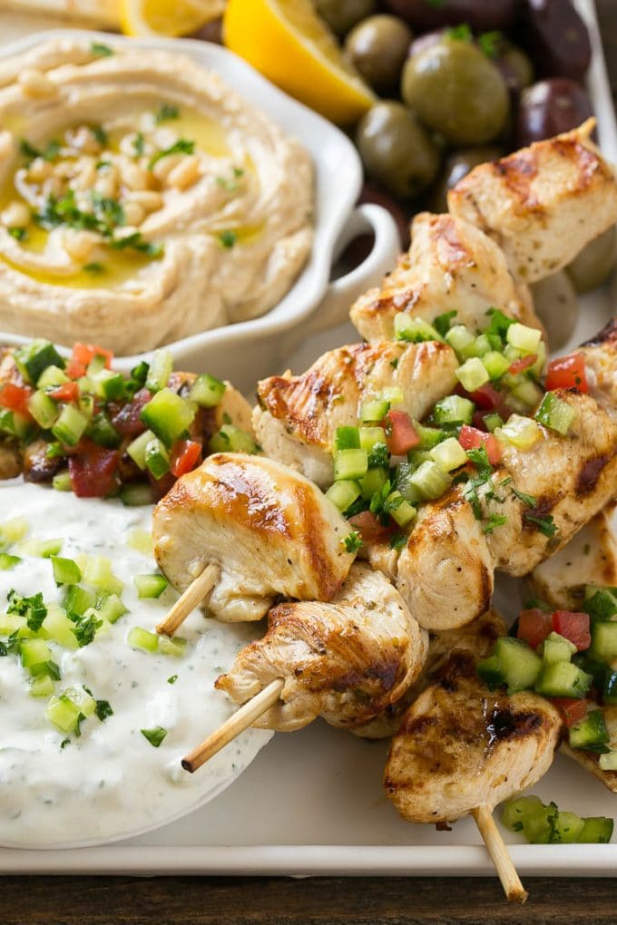Greek Chicken Souvlaki Recipe | Easy Greek Chicken | Chicken Souvlaki | Greek Chicken Skewers | Easy Greek Chicken Skewers #chicken #kabobs #glutenfree #keto #lowcarb #grilling #jonep A tray of chicken souvlaki kabobs with olives, hummus, pita bread and yogurt sauce.