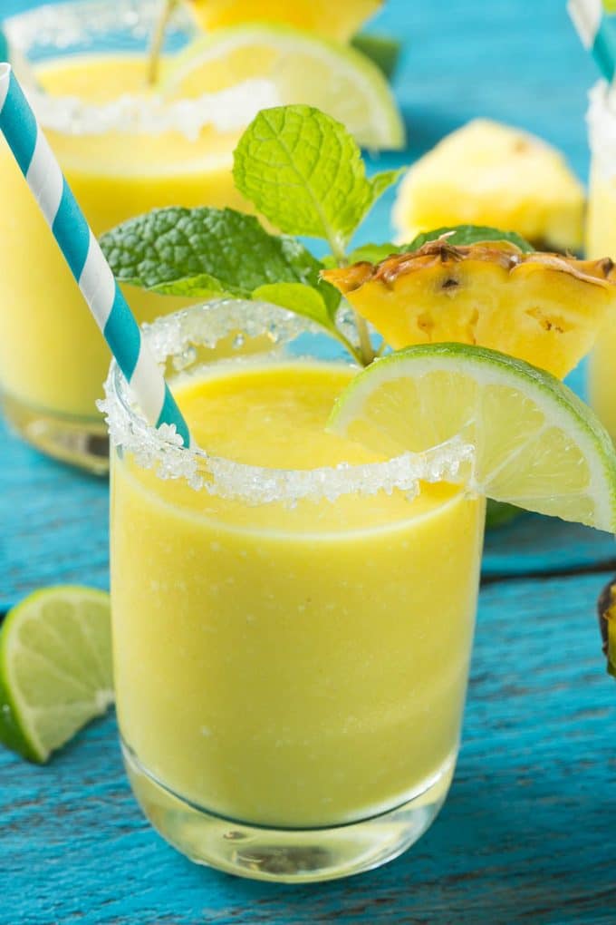Pineapple Coconut Smoothie Recipe |Tropical Smoothie | Pineapple Smoothie #smoothie #pineapple #coconut #healthy #cleaneating #jonep A cup of pineapple smoothie with garnishes of lime, pineapple wedges and mint sprigs.