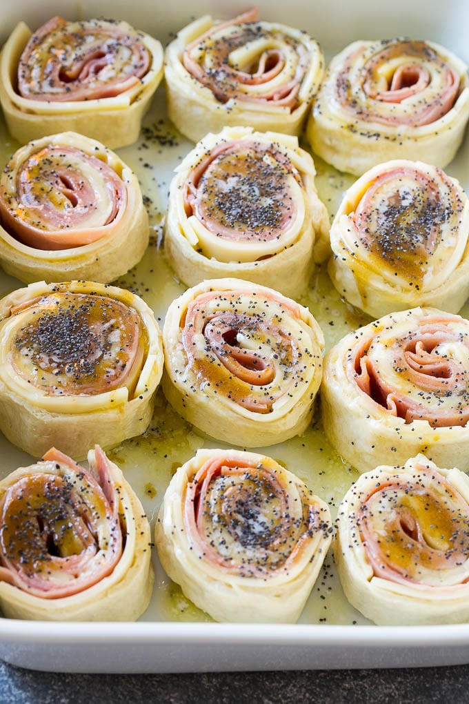 Ham and Cheese Pinwheels Recipe | Ham and Cheese Roll Ups | Ham and Cheese Rolls | Ham and Cheese Bread Rolls #ham #cheese #appetizer #pinwheels #jonep Dough rolled up with ham and cheese and topped with a poppy seed butter in a ceramic dish.
