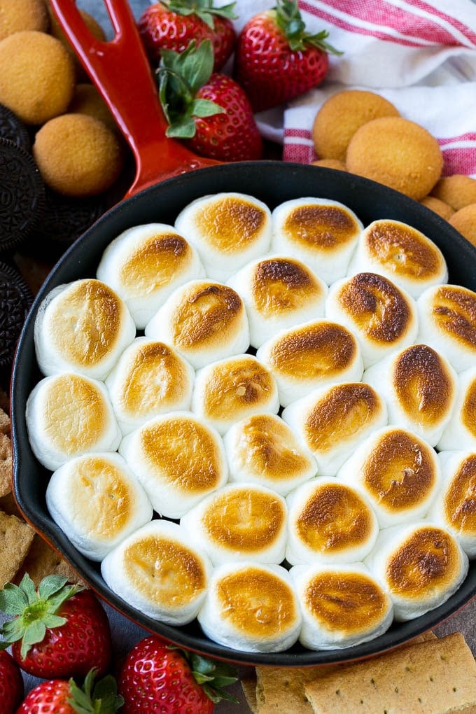 S'mores Dip Recipe | S'mores Skillet | Baked S'mores #smores #dip #chocolate #marshmallow #dessert #jonep A skillet of s'mores dip with melted chocolate chips and toasted marshmallows, surrounded by graham crackers, cookies and strawberries.