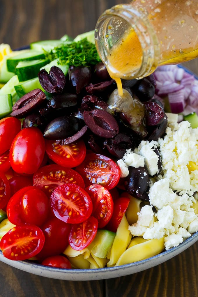 Greek Tortellini Salad Recipe | Pasta Salad | Tortellini Salad | Greek Salad #greek #tortellini #pasta #salad #cucumbers #olives #dinner #jonep Salad dressing being poured over a bowl of tortellini and vegetables.