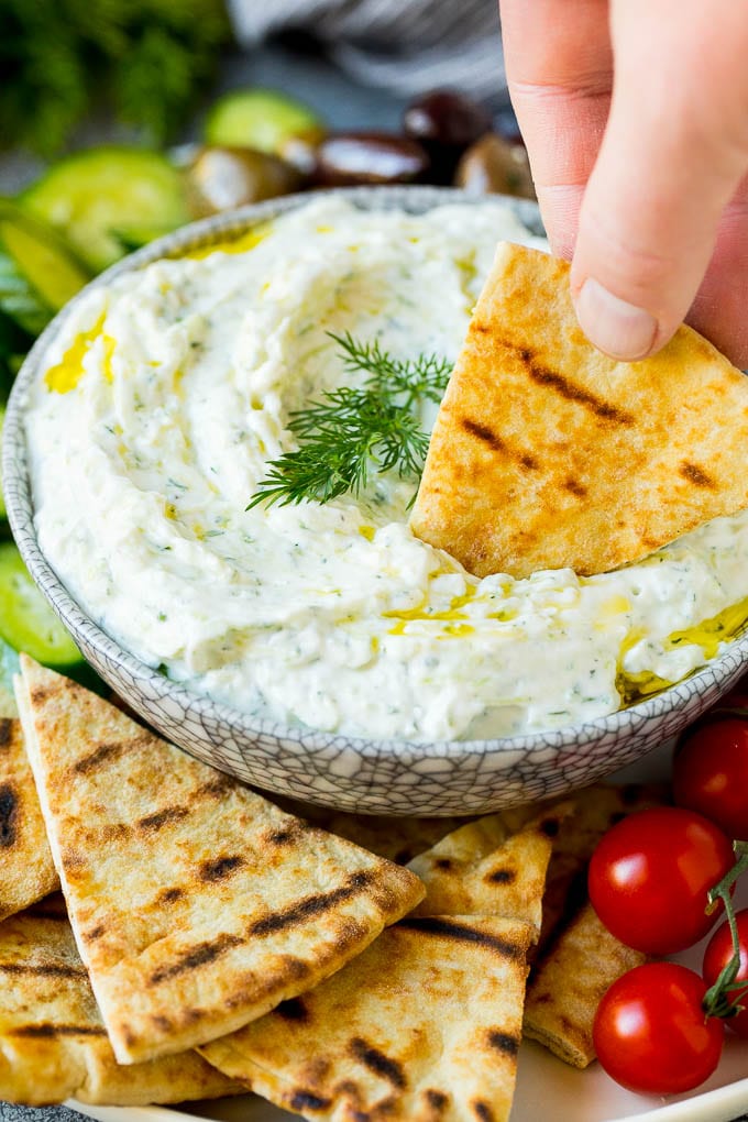 The best homemade tzatziki sauce with cucumber and herbs mixed into Greek yogurt. #appetizer #jonep A hand dipping pita bread into a bowl of tzatziki.