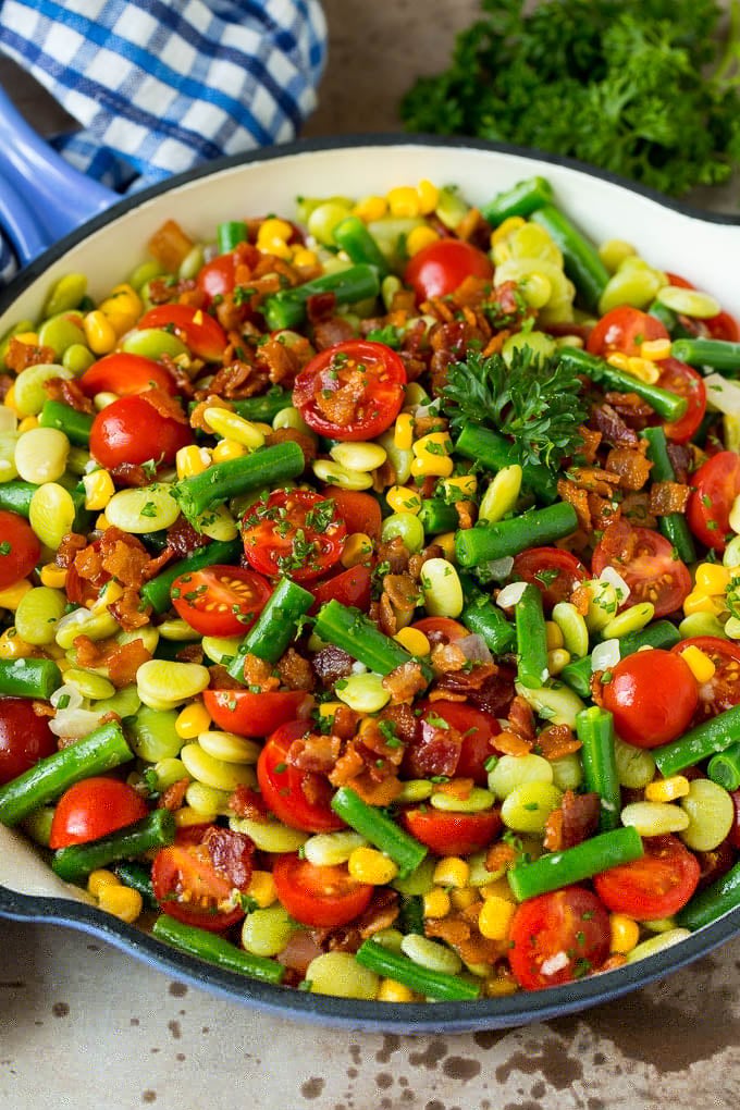 This succotash recipe is a combination of bacon, corn, tomatoes and lima beans, all cooked together to make a colorful and delicious side dish. A skillet of succotash topped with bacon and herbs.