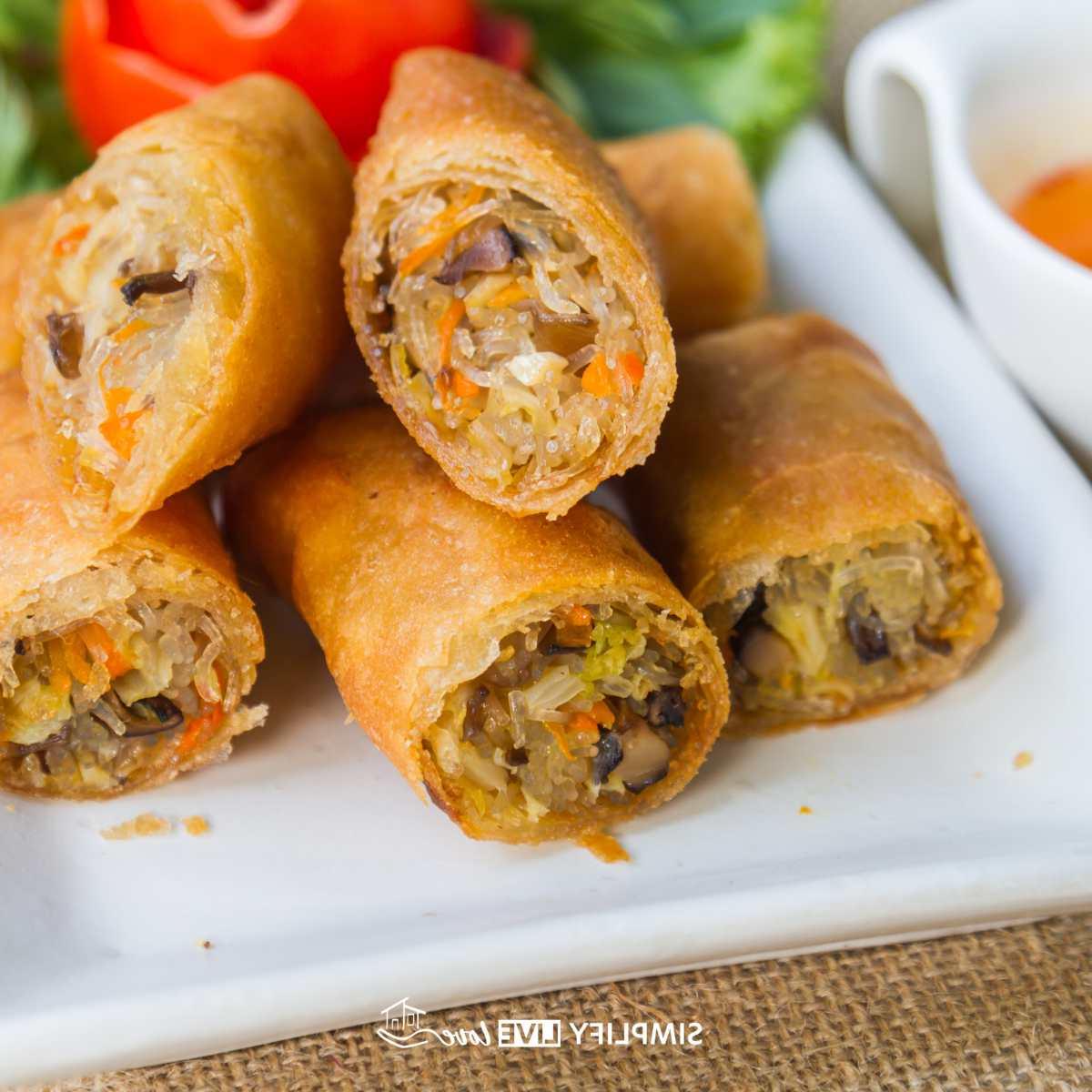 Maklike Resep vir Krokante Tuise Eierrolletjies met Boontjiespruite 2 tuisgemaakte eierrolletjies met kool en boontjiespruite op 'n wit bord met sous in die agtergrond