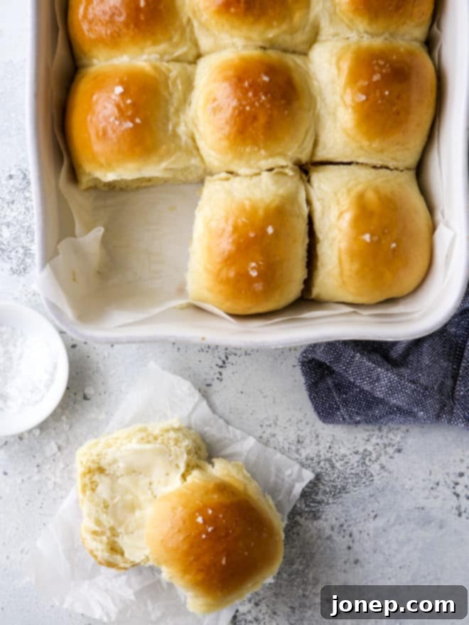 A close-up of soft white dinner rolls, some whole in the pan, and one sliced open with butter melting inside, highlighting their fluffy interior.