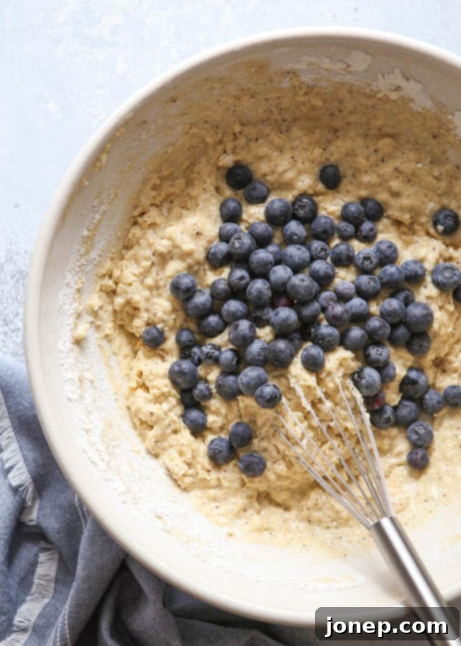 A close-up shot of homemade blueberry muffin batter, showing fresh blueberries gently folded into the mixture in a mixing bowl.