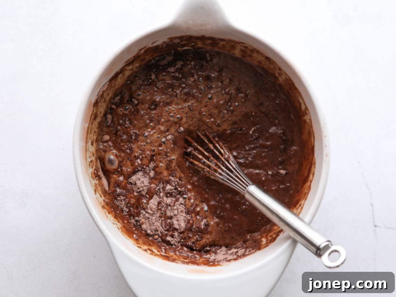 Close-up shot of rich, dark chocolate cupcake batter being gently mixed in a large white ceramic bowl, illustrating its smooth consistency with a few minor lumps.