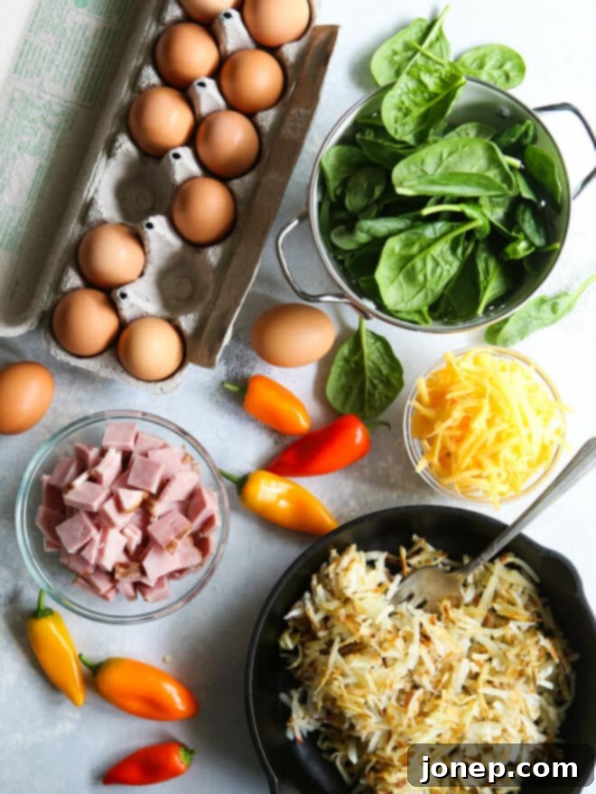 Ingredients for hash brown frittata laid out on a white surface, including peppers, spinach, ham, eggs, and cheese.