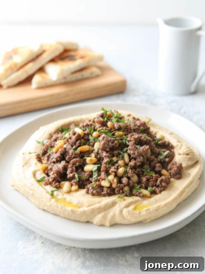 Overhead shot of a platter of creamy hummus piled high with savory spiced ground beef and golden toasted pine nuts, garnished with fresh parsley and mint, ready to be served.