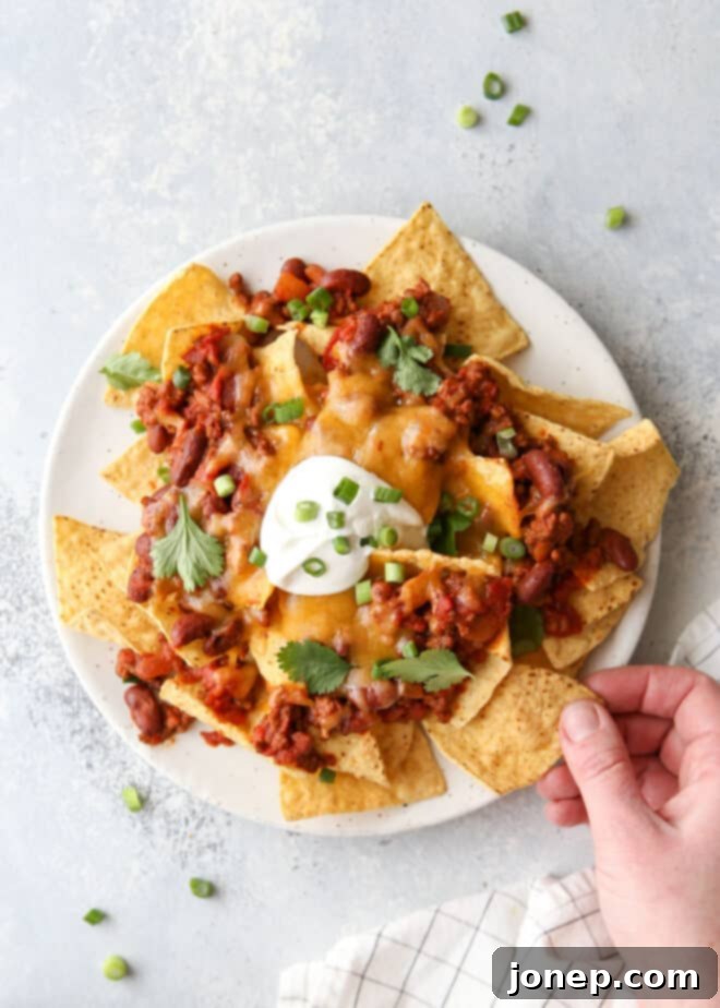 Ultimate Chili Cheese Nachos 6 A hand reaching for a chip from a platter of fully loaded chili cheese nachos
