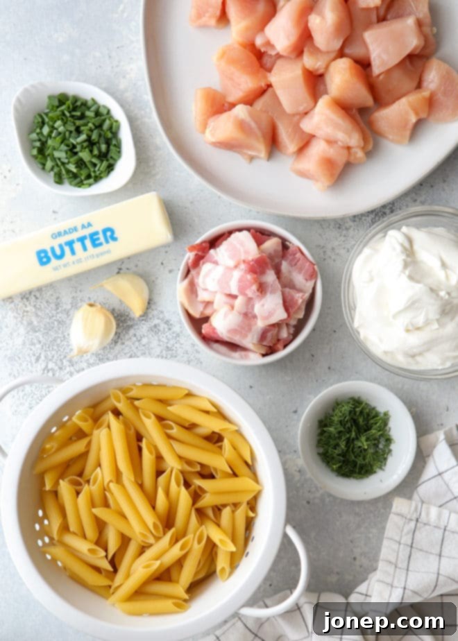 Close-up shot of the raw ingredients for Chicken Bacon Ranch Pasta, including penne pasta, cubed chicken breast, chopped bacon, fresh dill, chives, garlic, butter, and sour cream, neatly arranged on a wooden board.