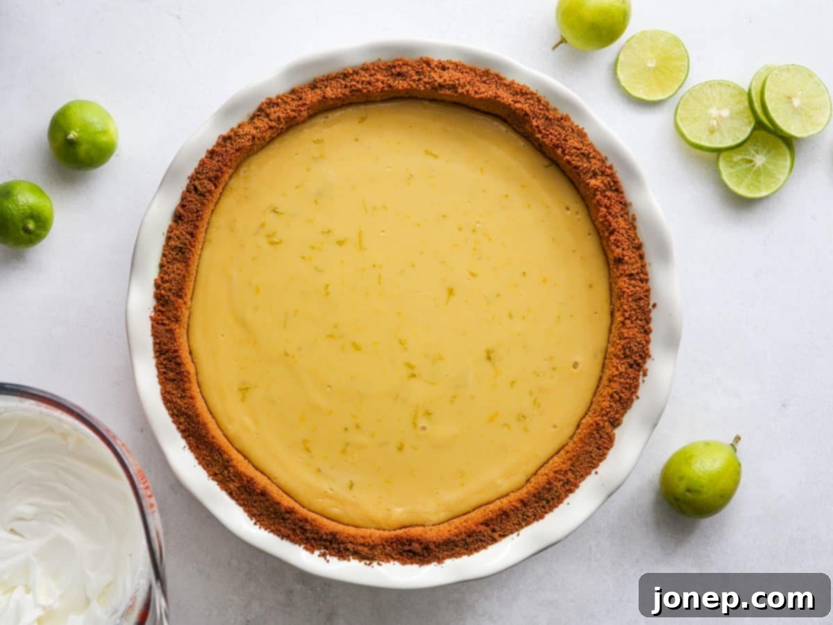A freshly baked Key Lime Pie cooling in a pie plate, surrounded by ingredients like whipped cream in a bowl and sliced limes, ready for topping and chilling.