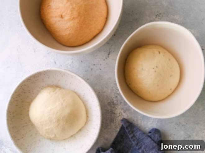 Three separate bowls, each containing a different type of risen dough – traditional white, honey wheat, and herb – ready for shaping, demonstrating variety.