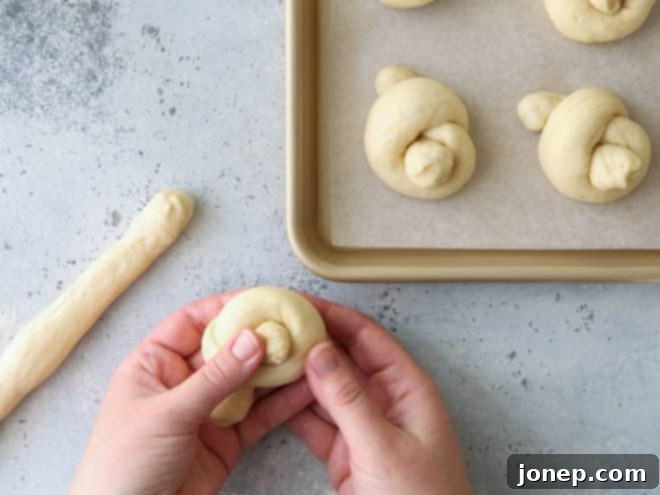 Mastering Dinner Roll Shapes 7 Hands skillfully tying a portion of white dinner roll dough into an elegant knot on a clean work surface.
