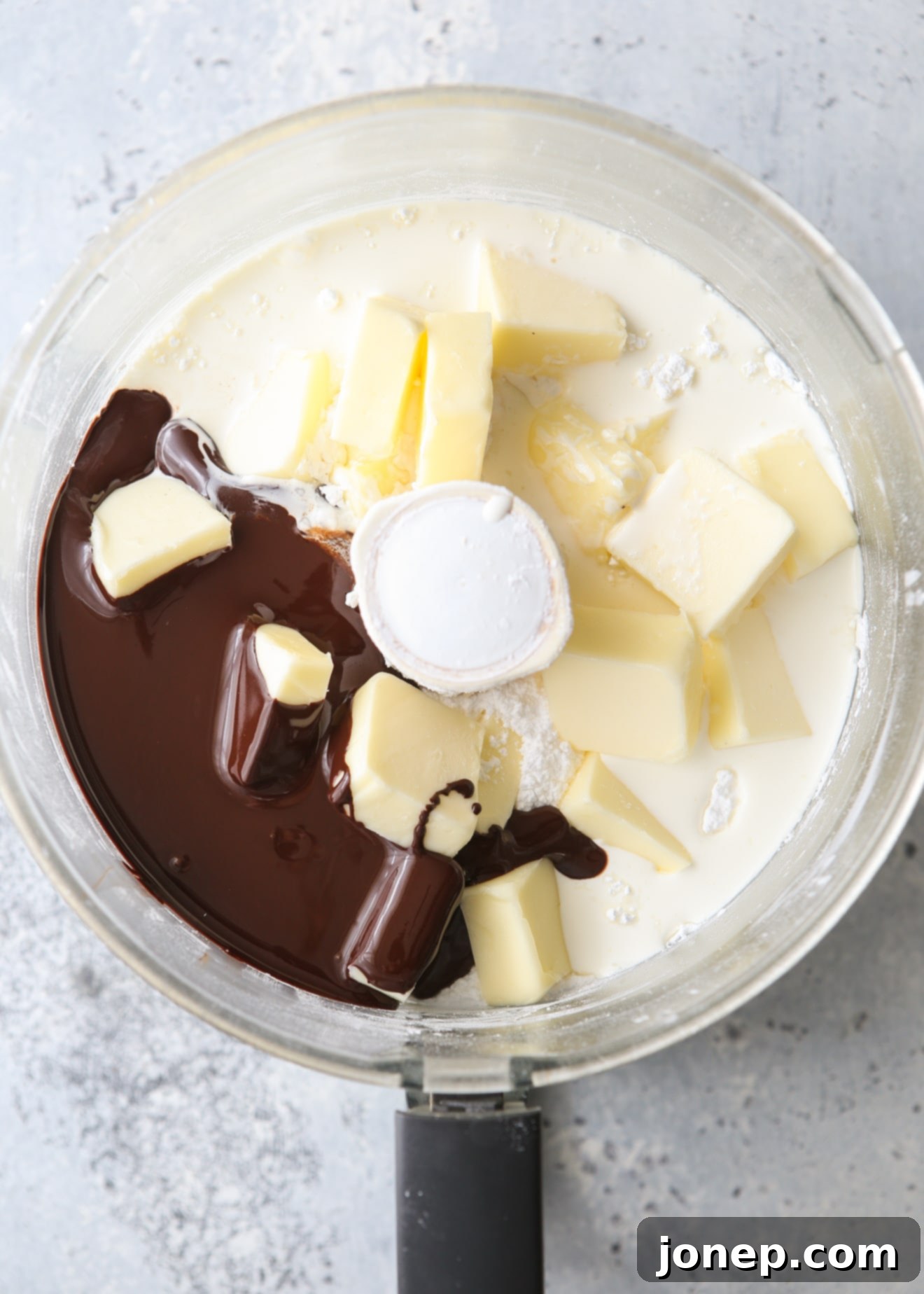 All the ingredients for fudge buttercream, including melted chocolate, powdered sugar, butter, and vanilla, placed in a food processor bowl before blending