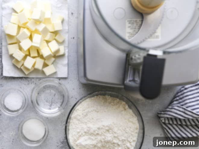 Effortless Food Processor Pie Crust 3 Essential ingredients for pie crust, including flour, butter, and ice water, next to a food processor.