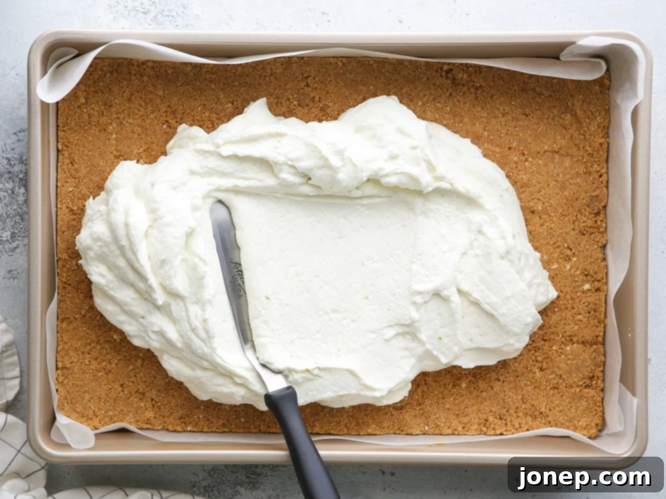 A hand spreading the creamy, light green cheesecake filling evenly over the chilled graham cracker crust in a baking pan.